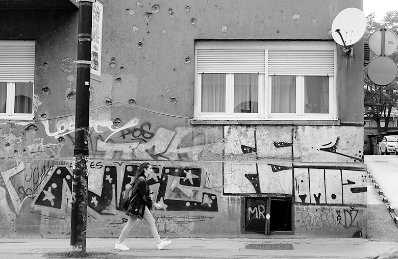 Eine Gebäudefront samt Fernsehantenne. Der untere Teil der Wand ist von Graffiti überzogen, der Putz bröckelt teilweise schon ab. Vor dem Gebäude läuft ein Mädchen auf dem Bürgersteig und ein großer Schwarzer Mast ragt aus dem Boden. Neben dem Haus ist ein Teil einer Straße zu sehen, auf der ein Auto fährt.