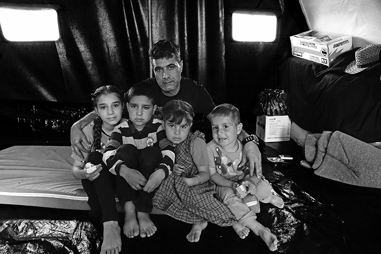 Shahed sitzt mit drei jüngeren Kindern eng aneinander gedrängt auf einer Matratze. Ein Mann mit kurzem, dunklem Haar und ernstem Blick, sitzt nach vorn gebeugt schützend hinter ihnen. Seine Arme sind ausgebreitet und die Hände liegen auf den Schultern der beiden Kinder, die am Rand sitzen.