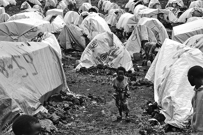Ein provisorisch wirkendes Flüchtlingslager. Niedrige, mit Steinen befestigte Zelte aus weißen Planen reihen sich aneinander. In der Mitte des Fotos läuft ein junges Mädchen bergauf auf die Kamera zu. Sie trägt ein Baby auf ihrem Rücken.